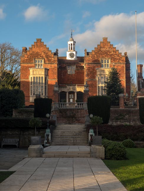 A historic red brick building with Gothic architectural features, including stepped gables and tall, narrow windows, set against a blue sky with scattered clouds. In the foreground, there is a neatly