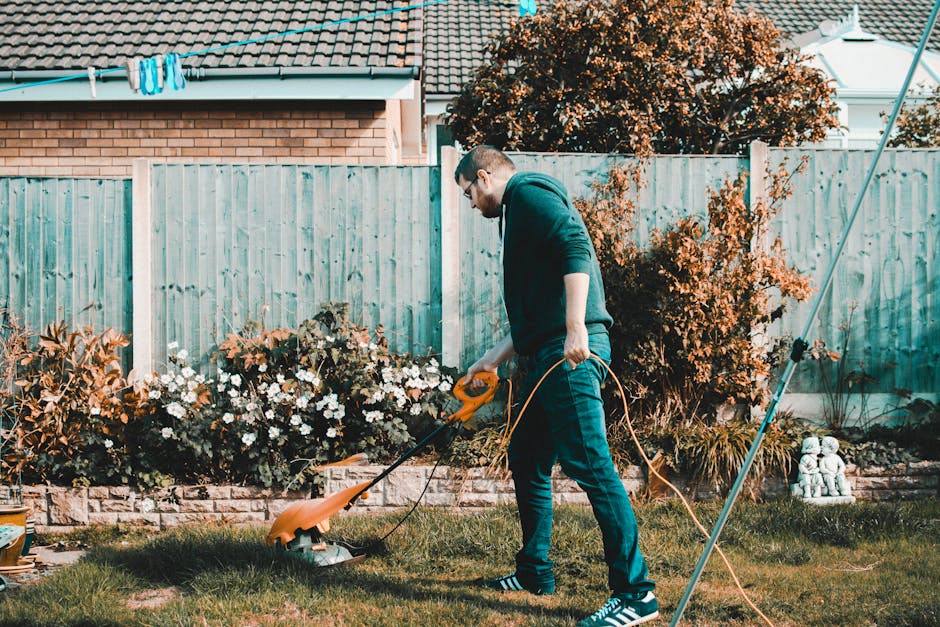 A man in casual clothing, including a dark hoodie and blue jeans, is performing garden maintenance in a backyard, using an electric string trimmer to trim grass and weeds along a lawn border. The gard